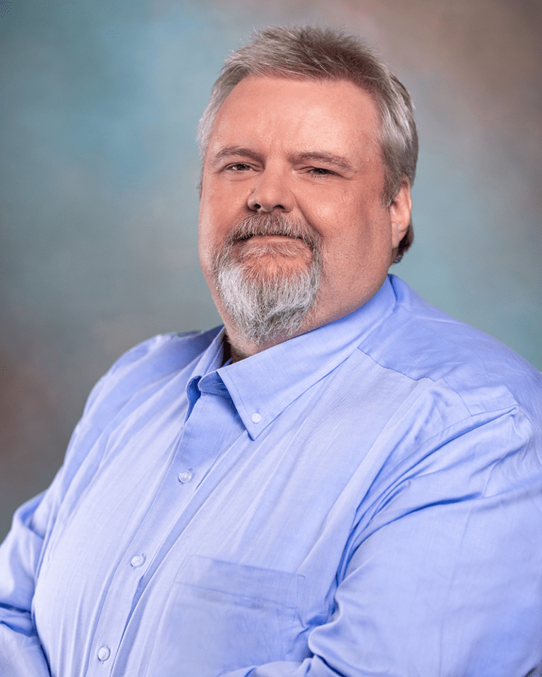 A man with a beard and mustache wearing a light blue button-up shirt poses with a slight smile against a blurred background.