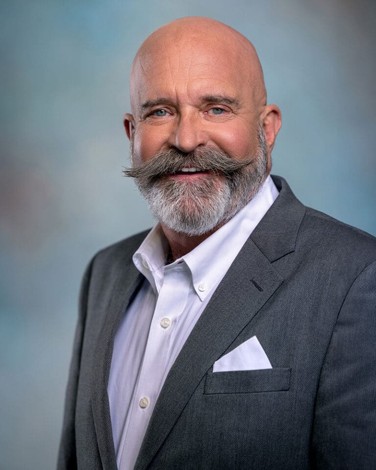 A bald man with a large, curled mustache and beard is wearing a white dress shirt and a gray suit jacket. He is smiling against a blurred background.