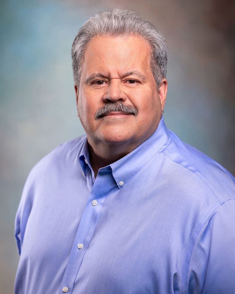 A man with gray hair and a mustache wearing a light blue button-up shirt poses against a gradient background.