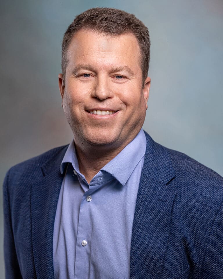 A man in a blue blazer and light blue shirt smiles with a neutral background.