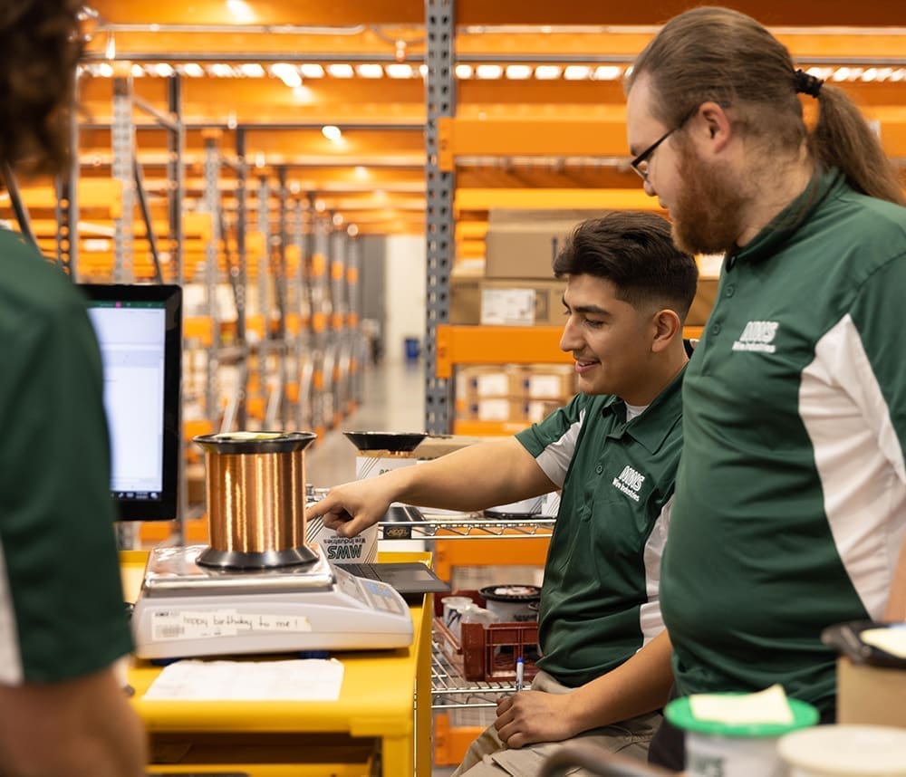 Three people in green shirts are working in a warehouse, focusing on a spool of copper wire on a scale. Shelves and boxes are visible in the background.