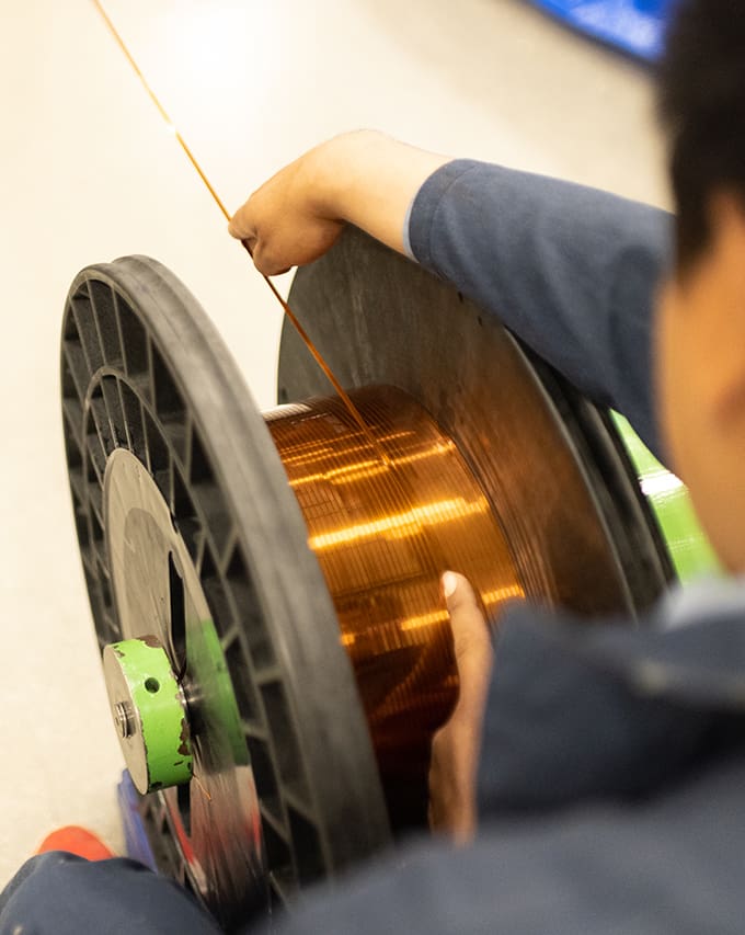 A person winds flat wire onto a large black spool, holding the wire with one hand and guiding it with the other.