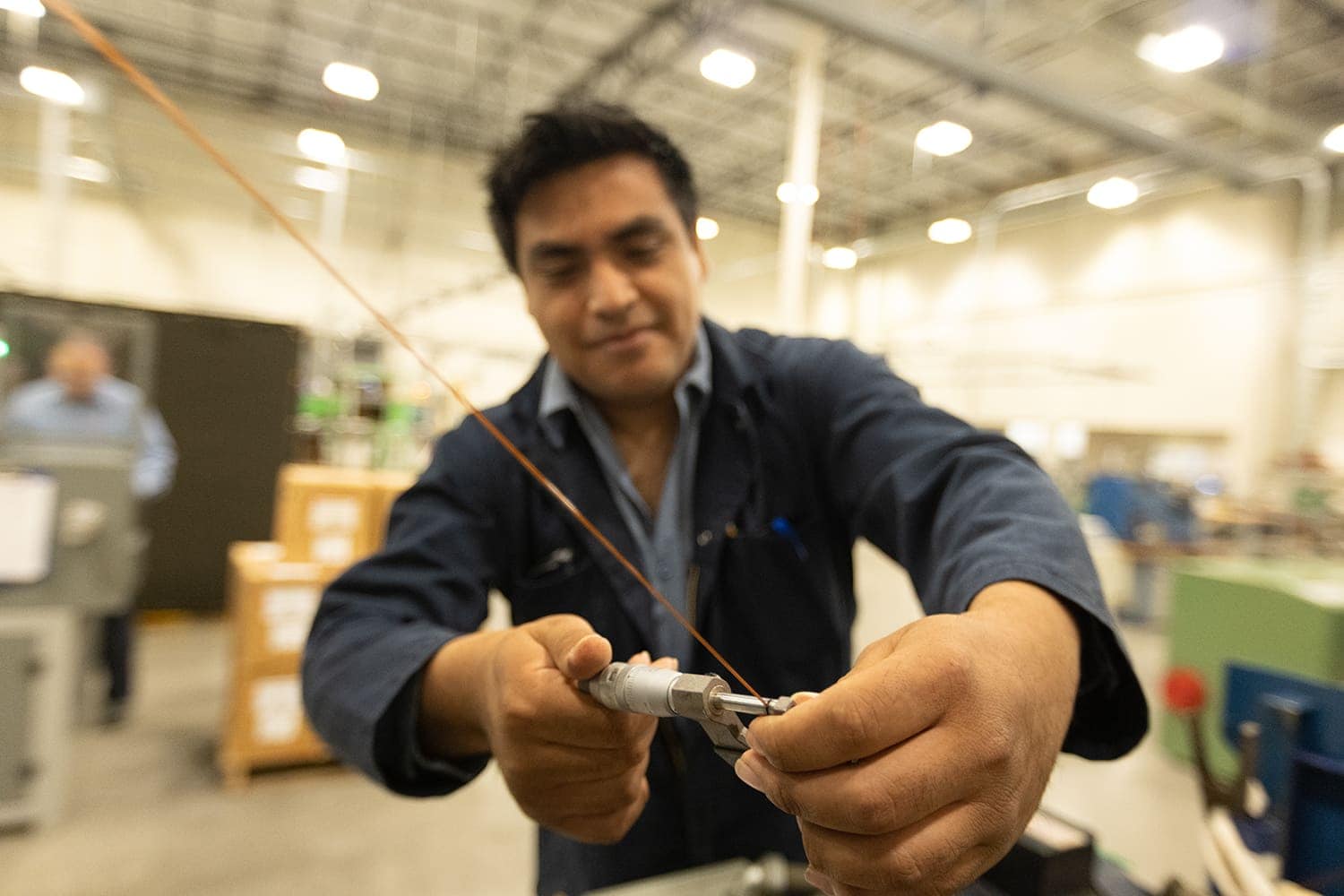 A person in work attire measures a flat wire with a caliper inside a factory or industrial setting