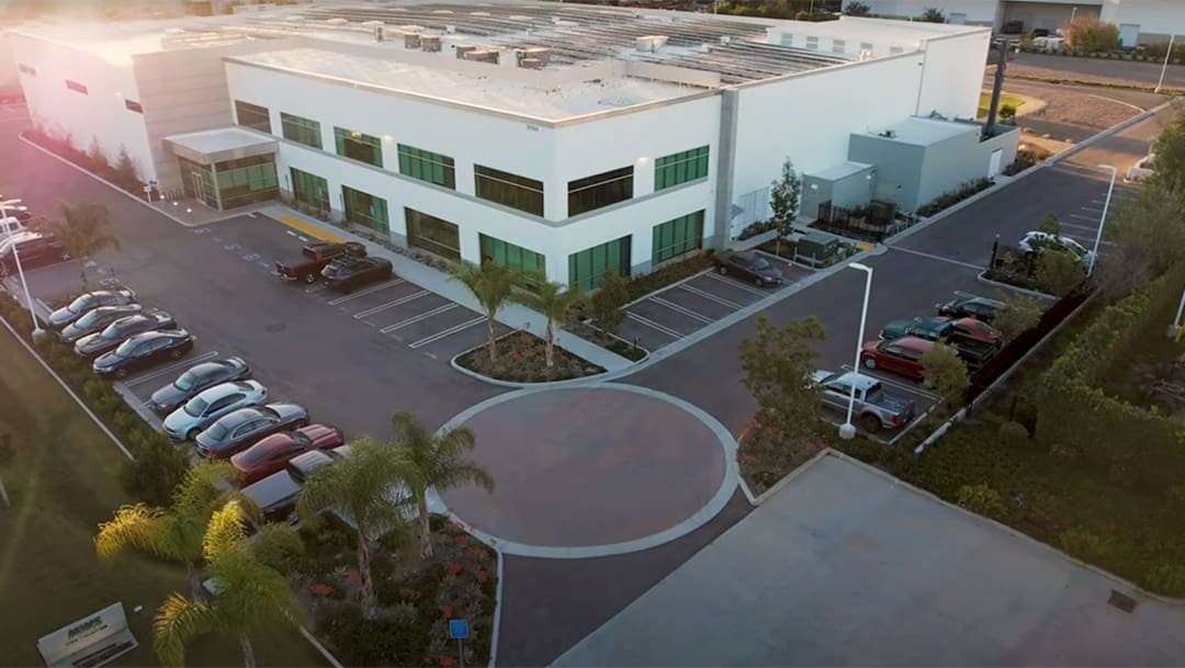 Aerial view of a modern two-story office building with adjacent parking lot, several cars parked, and landscaped greenery surrounding the property.