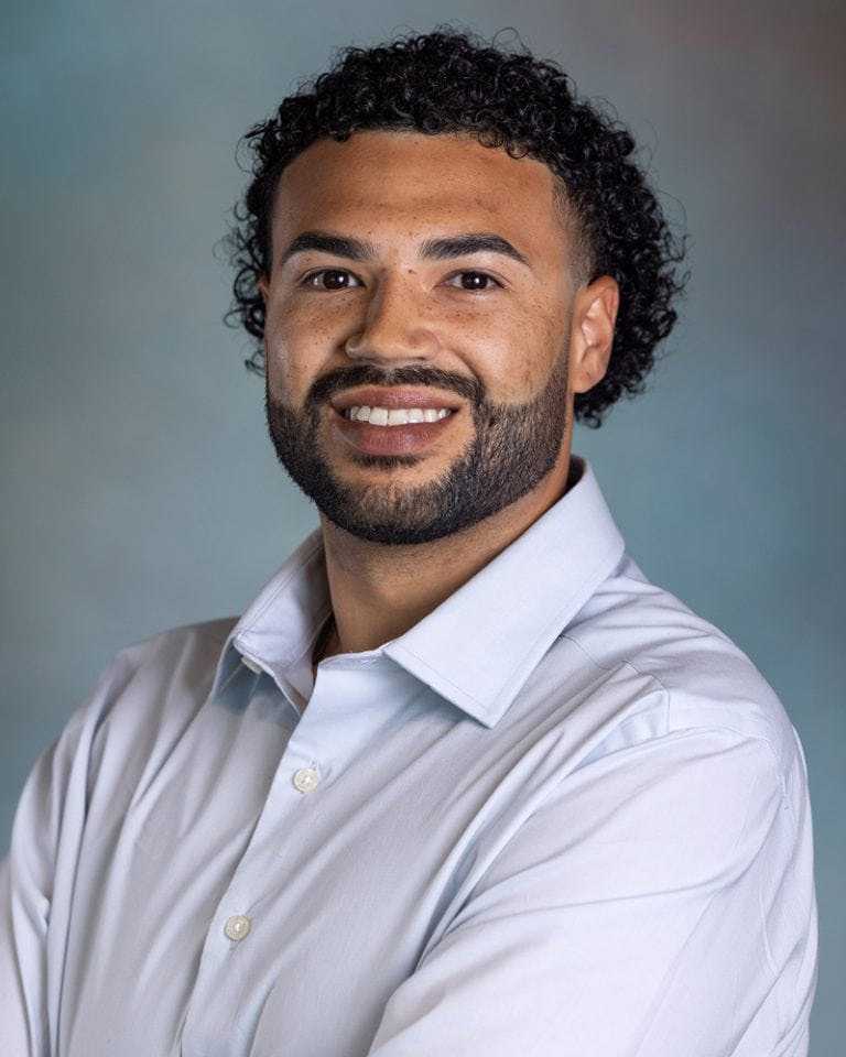 Headshot of Javon Bell with curly hair and beard, wearing a white shirt, smiling confidently for a professional profile.