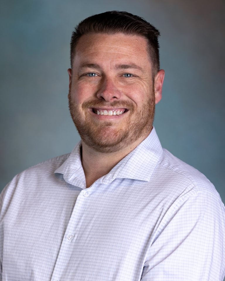 Close-up portrait of Keith Zvorak, smiling man with short brown hair and beard, wearing a light blue checkered shirt, against a neutral background.