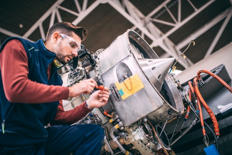 A technician in safety glasses and a blue vest inspects and repairs satellite or spacecraft equipment inside a high-tech facility.