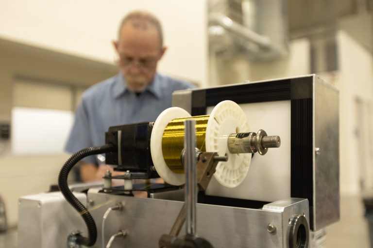Close-up of custom wire production machinery with spools of gold wire and a technician in the background at MWS Wire.